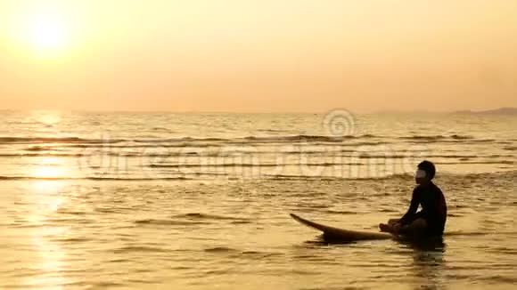 4K冲浪者的剪影放松坐在冲浪板在海面上的日落热带海滩体育和娱乐视频的预览图