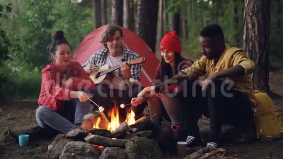 快乐的徒步旅行者正在火上煮棉花糖唱歌而帅哥在度假时弹吉他视频的预览图