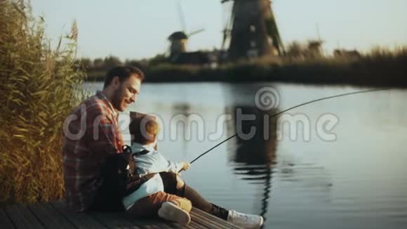 欧洲父子坐在湖边码头上男孩握着一只手工制作的渔具家庭关系美满4K视频的预览图