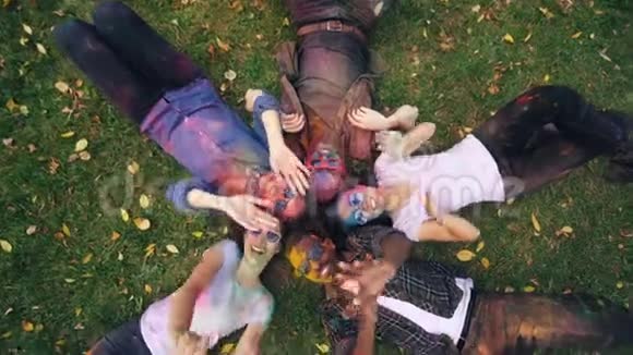 欢乐的女孩和男孩躺在公园的草地上他们的脸和衣服上涂满了多色的油漆人们都喜欢视频的预览图