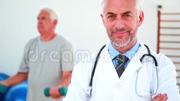 微笑医生看着摄像机病人在后台锻炼视频的预览图