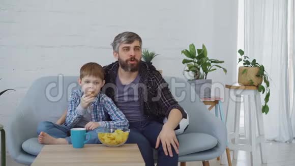 足球迷们留着胡子的男人和他的小孩在家里看电视庆祝进球和吃薯片视频的预览图
