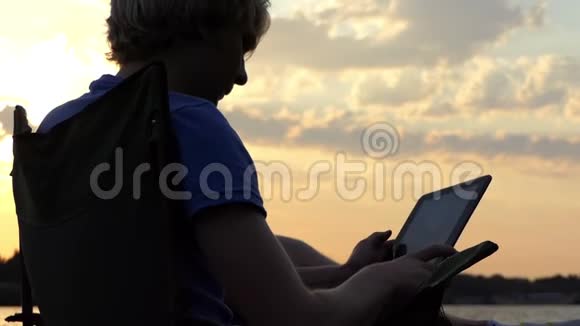 年轻的博客作者坐在椅子上看着他在斯隆莫日落时的平板电脑视频的预览图