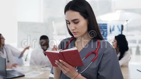 重点突出的白种人女医生在笔记本上做一些笔记而工作人员小组则在视频的预览图
