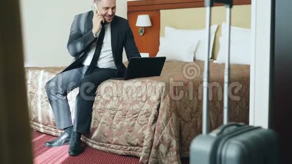 坐在酒店房间的床上忙碌的商人穿着西装在笔记本电脑上工作在手机上说话视频的预览图
