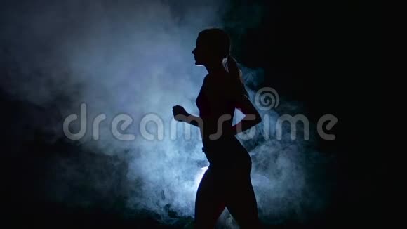 运动健身女孩在黑色背景上奔跑被烟雾中的聚光灯照亮剪影慢动作视频的预览图