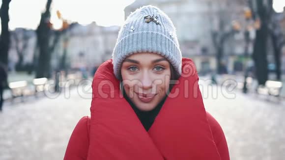 一个戴着灰色帽子和红色外套的漂亮女孩在圣诞节装饰前站在街上微笑的肖像视频的预览图