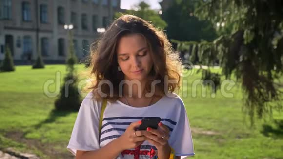年轻的白种人女孩站在公园里正在使用智能手机思考大学背景视频的预览图