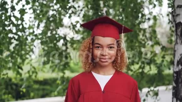 穿着红色长袍和灰泥板的漂亮非裔美国女毕业生站在户外微笑着的肖像视频的预览图