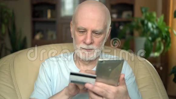 英俊的微笑老人坐在家里的椅子上用信用卡在线购买智能手机视频的预览图