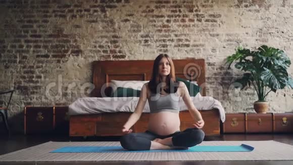 运动孕妇正在家中练习瑜伽坐在卧室地板上的垫子上举手把手掌放在一起视频的预览图