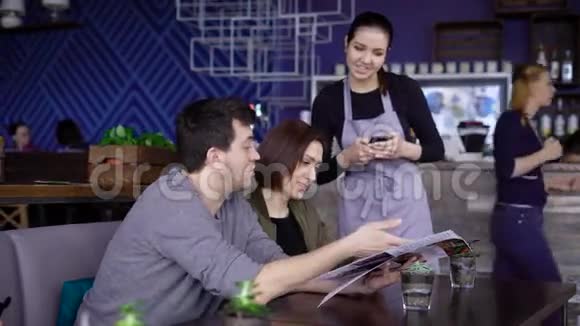 年轻的微笑女服务员站在幸福夫妇的桌子附近的咖啡馆里把他们的订单放进数字平板电脑里男子视频的预览图