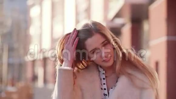 一位年轻漂亮的金发女郎微笑着对着镜头触摸她的头发在市中心醒来的肖像视频的预览图