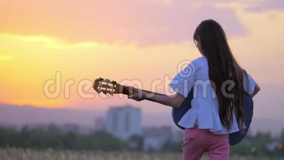 日落时美丽的风景有趣的小女孩音乐家弹吉他走在户外蒲公英田野远离城镇视频的预览图