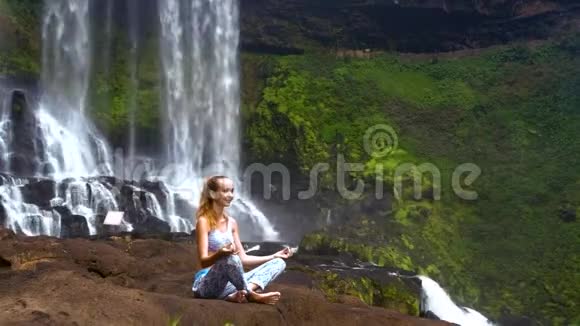 女孩在瀑布的大石头上摆着普拉纳亚摩瑜伽姿势视频的预览图
