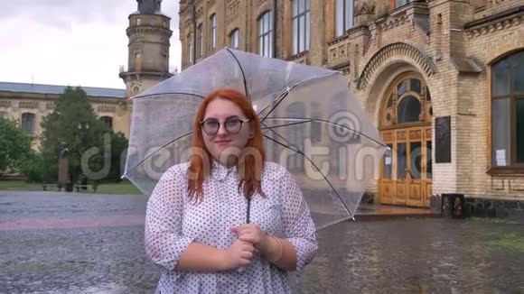 戴眼镜的胖姜女站在雨天的公园里撑着伞看着镜头微笑着视频的预览图