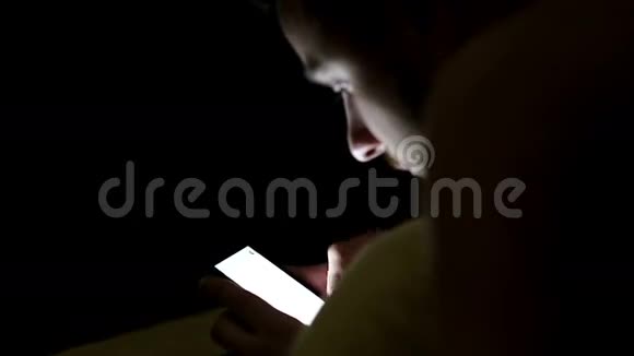 一个年轻人晚上在毯子下面的床上使用智能手机视频的预览图