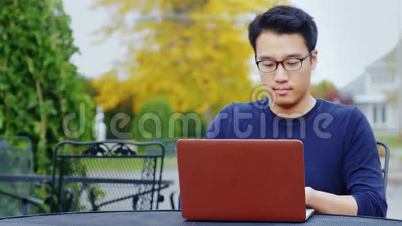 一个年轻的亚洲男人用笔记本电脑工作在典型的美国小镇上坐在户外视频的预览图