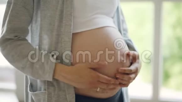 期待的母亲正在抚摸她的大婴儿肿块站在室内背景是大窗户快乐的怀孕人们视频的预览图