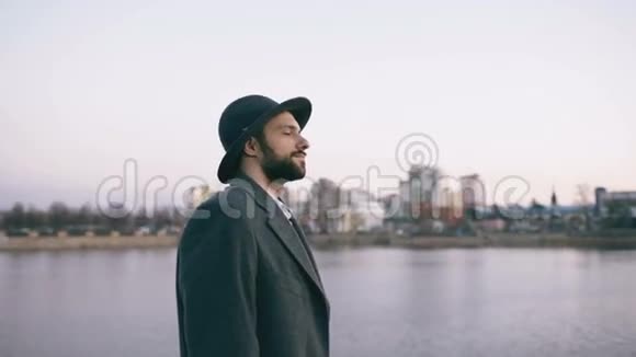 站在河边时戴着帽子和外套的留着胡子的年轻游客正在观看城市景观和白日梦视频的预览图