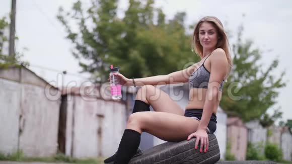 一位年轻漂亮的运动型金发女郎穿着上衣和短裤坐在轮胎上喝着瓶子里的水视频的预览图