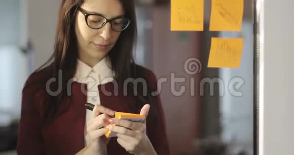漂亮的女商人戴着眼镜在办公室的玻璃上写着贴纸和胶水上的信息视频的预览图