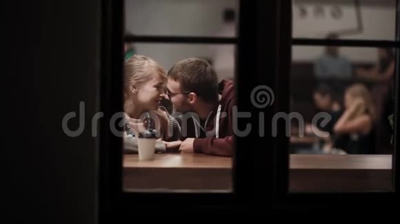 通过窗口查看年轻漂亮的夫妇坐在咖啡馆里聊天晚上在市中心的浪漫约会视频的预览图