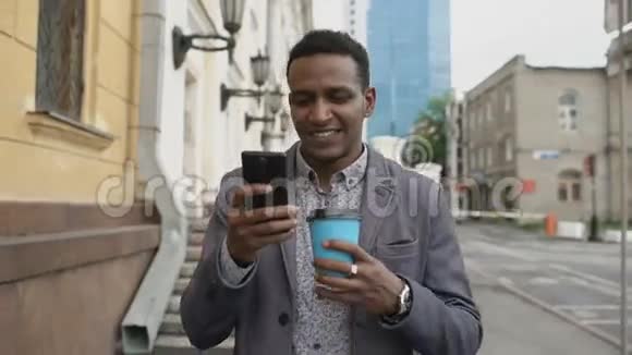 stadicam拍摄的年轻快乐商人使用智能手机在户外端着咖啡散步视频的预览图