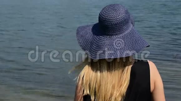 戴着帽子的女孩看着平静的水面直到她的头发在风中飞舞从后面看视频的预览图