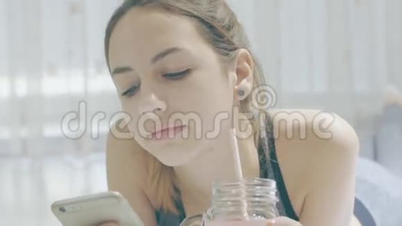 这名年轻女子正在喝冰沙并在智能手机上使用健身应用程序视频的预览图
