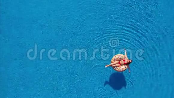 一位穿红色比基尼的女士躺在游泳池的甜甜圈上的空中景色视频的预览图