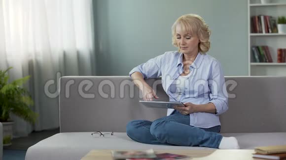 漂亮的金发老年女性坐在沙发上在平板电脑应用程序上滚动视频的预览图