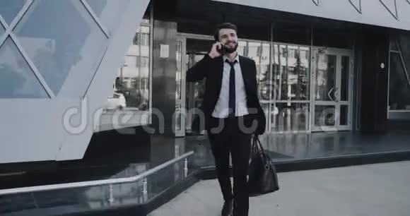 快乐的有魅力的年轻的事业的男人的来的出来的办公室的大楼的说话的电话的拿着的他的公文包视频的预览图