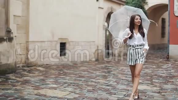 微笑着年轻的黑发女人穿着衣服带着雨伞沿着一个古老的城镇的街道散步在雨下散步视频的预览图