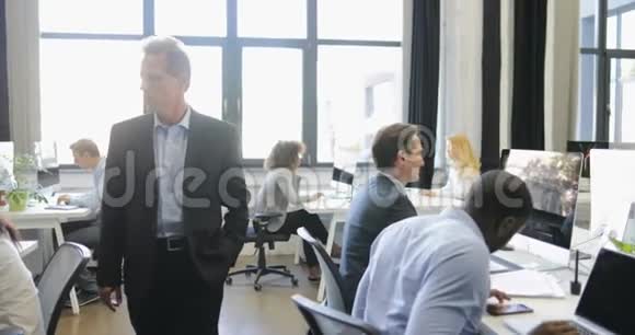 老板走在现代的开放式办公室而一群商业人士在电脑上工作领导看混合种族小组视频的预览图