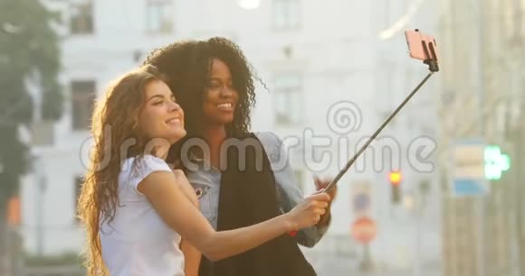 在街道上用自拍棒拍照的美丽微笑的多民族女朋友的横向视角视频的预览图