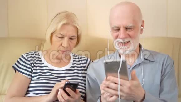 开朗好看的老两口坐在沙发上带耳机的智能手机听音乐视频的预览图