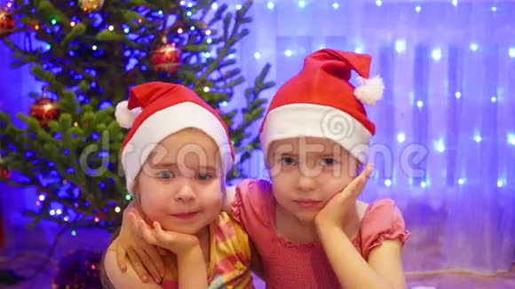两个可爱的女孩微笑着戴着圣诞帽对着镜头挥手背景圣诞杉木的灯光和花环视频的预览图