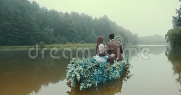 这对时尚的情侣坐着老式的树懒漂浮在浪漫的小船上沿着雾中的绿色草本植物装饰着视频的预览图
