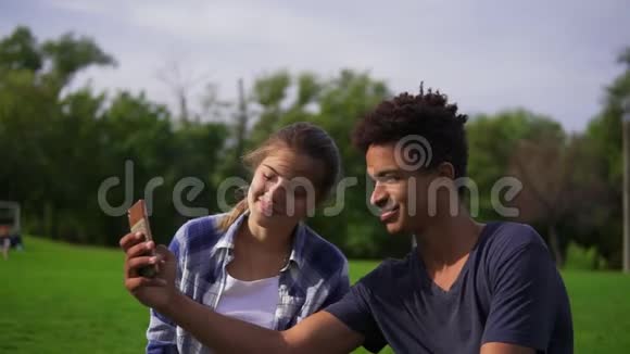 可爱的朋友在公园里休息坐在草地上看着智能手机笑着一对混血儿视频的预览图