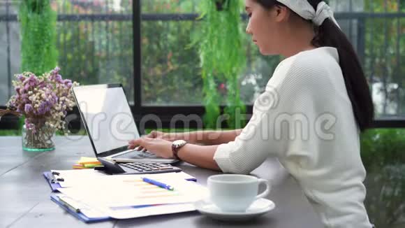 美丽的年轻压力亚洲妇女在笔记本电脑上工作而不高兴使用智能手机在客厅在家里视频的预览图