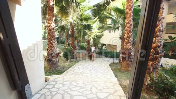 年轻的黑发女人穿着短裤太阳镜和高跟鞋带着手提箱在酒店的棕榈树之间假期视频的预览图