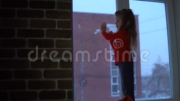 小可爱的女孩站在窗台上望着外面的雪城风景视频的预览图