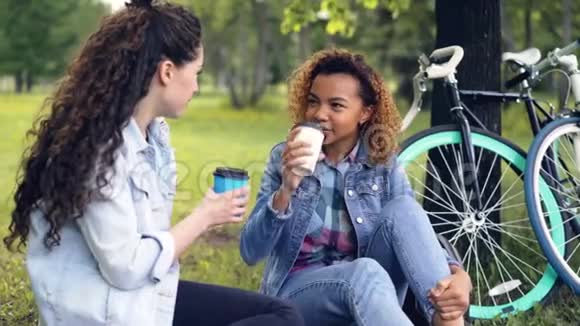 游客朋友们正在喝咖啡笑着坐在城市公园的草地上骑着自行车靠近树年轻有魅力视频的预览图