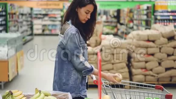 美丽的女人快乐的顾客在超市买水果选择香蕉和苹果把它们放进购物车视频的预览图