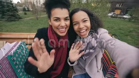 两个快乐的混血女人带着购物袋在视频电话中和SmartPone交谈坐在长椅上的年轻女孩视频的预览图