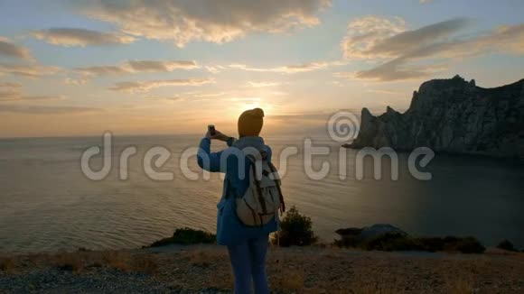 年轻迷人的女孩徒步旅行在智能手机上拍照在海边的海角上拍摄美丽的日落达到了目标视频的预览图