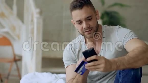 有魅力的年轻人带着智能手机和信用卡在网上购物坐在家里的床上视频的预览图