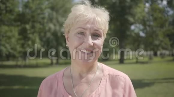 一位白发苍苍的老妇人站在夏季公园里微笑着看着摄像机户外休闲成熟的白种人女士休息视频的预览图