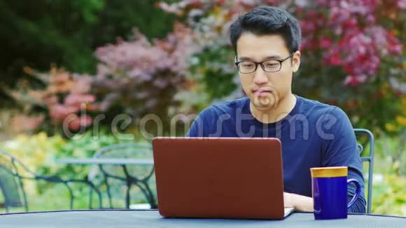 戴眼镜的亚洲人用笔记本电脑工作坐在花园或咖啡馆的夏日露台上视频的预览图